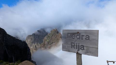 Wooden sign 'Pedra Rija' against a mountainous cloud-covered backdrop.