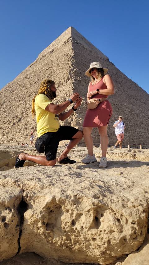 A man kneels in front of a woman near a pyramid with tourists around.