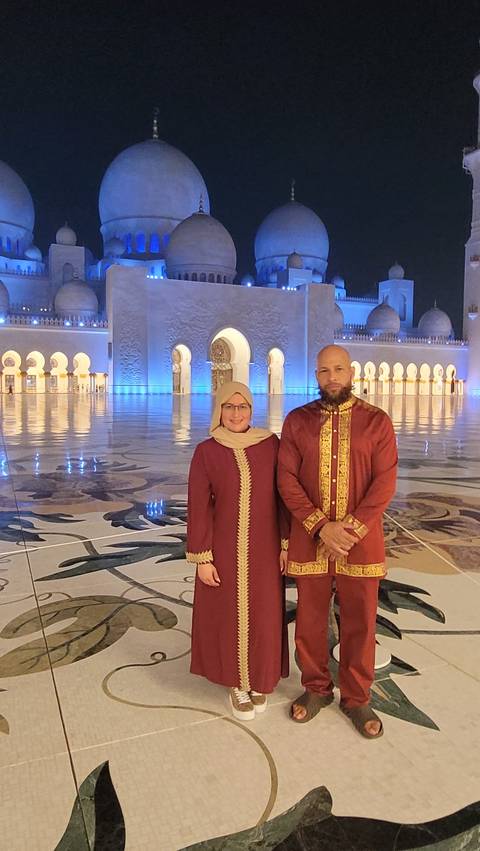 Two people in traditional attire with an ornate mosque backdrop illuminated at night.