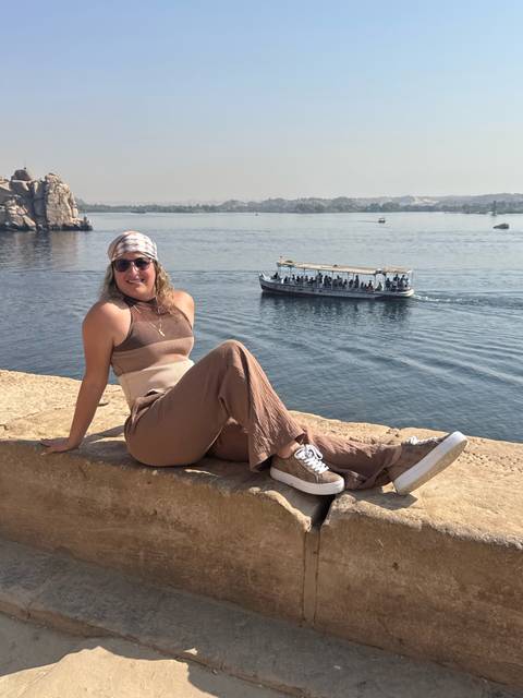 A woman sitting on a ledge overlooking a river with a boat passing.