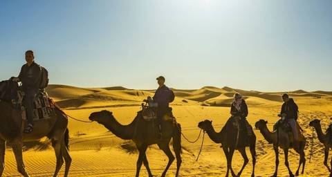People riding camels in the desert under a bright sun.