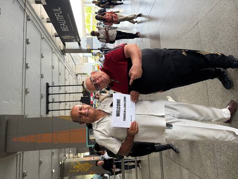 Two men posing with a welcome sign at an airport.