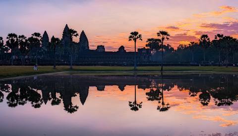 Sunset view of Angkor Wat reflected in a pond with palm trees.