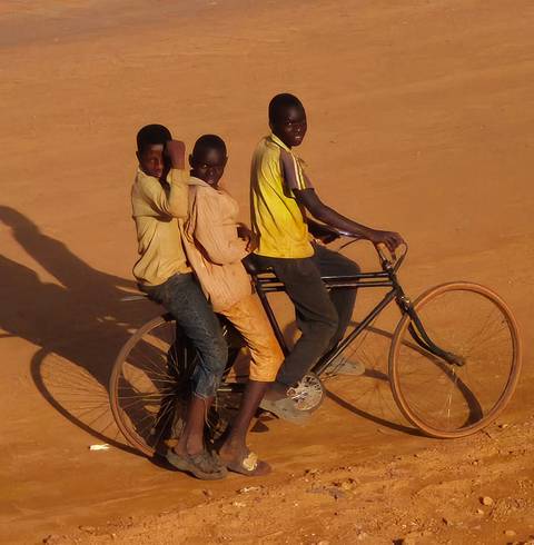 Three people riding a bicycle on a dirt road.