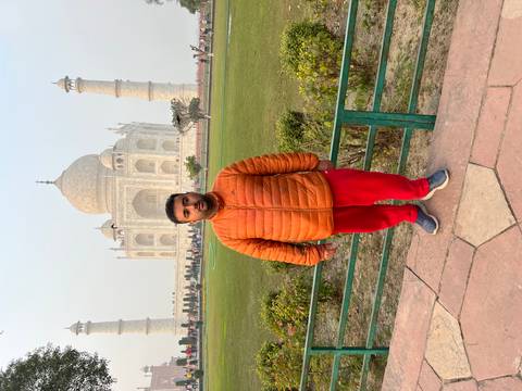Person posing in front of the Taj Mahal with greenery in the foreground.