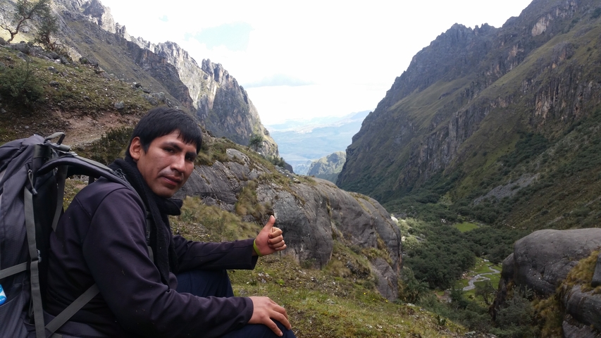 Guide sitting on a mountainside trail giving a thumbs-up with dramatic green valley stretching behind.