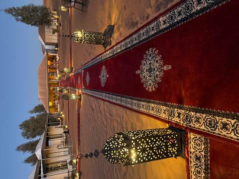 Pathway lined with elegant lanterns and red carpets leading to a tent in the desert at twilight.