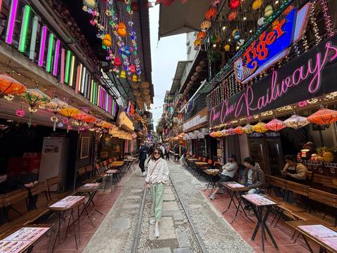 A vibrant street with colorful lanterns and people enjoying outdoor cafes beside a railway track.