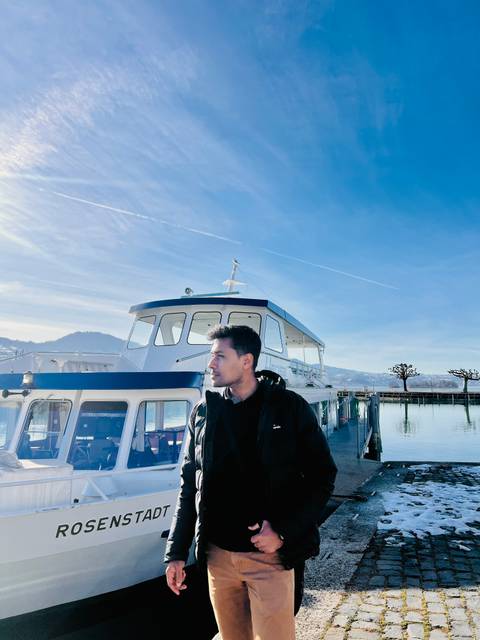 Person standing by a boat on a sunny day with mountains in background.