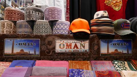 Colorful market stall stacked with patterned Omani caps, hats, souvenir licence plates and folded scarves.