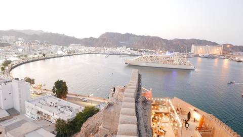 Panoramic evening view over Muscat harbour with a large white cruise ship anchored in the calm bay framed by jagged mountains.