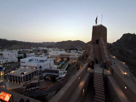 Stone stairway up to a lookout tower on a rocky ridge above Muscat’s white government buildings at dusk.