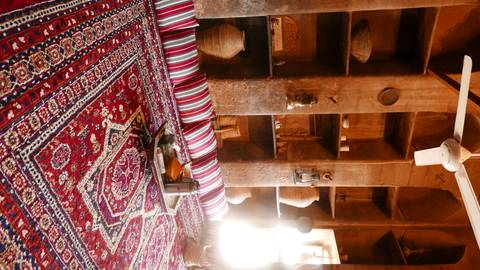 Cozy traditional majlis room with striped cushions, patterned red carpet and shelves filled with earthenware and lanterns.