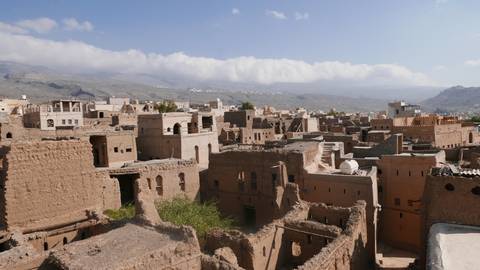 Panoramic view of crumbling mud-brick houses of an old Omani town with mountains and scattered clouds beyond.