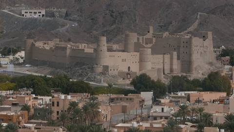 Distant view of massive sand-coloured Bahla Fort rising above palm-strewn town and rocky hills.
