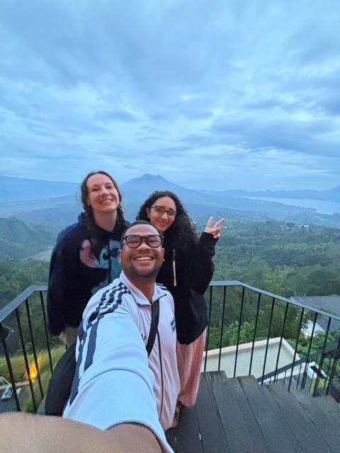 Three friends taking a selfie with a scenic view of hills and a lake.