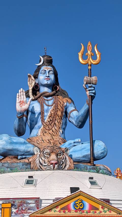 Tall blue statue of the Hindu god Shiva against a vivid blue sky, richly detailed with ornaments.