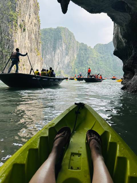 Kayaks and small tour boats filled with life-jacketed visitors paddle through emerald waters beneath towering karst cliffs and a cave arch.