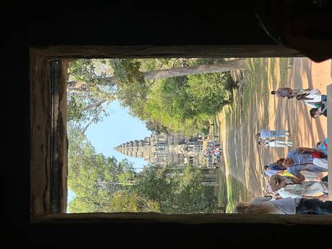 View from dark doorway toward crowds walking a forested avenue to an Angkor temple tower.