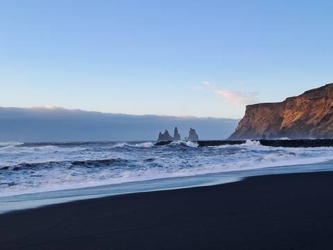 Waves crash on black sand beach with dramatic Reynisdrangar sea stacks and cliff at dusk.
