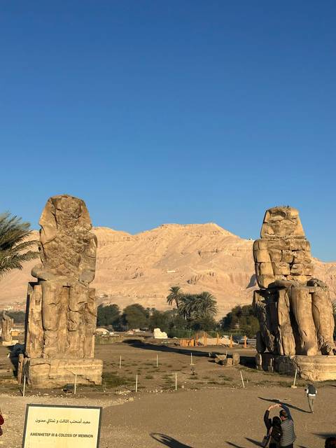 Two ancient stone statues facing a desert mountain backdrop under a clear blue sky