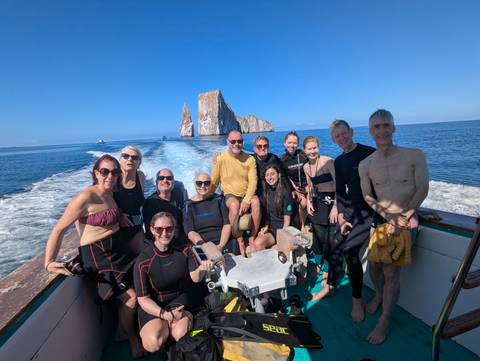 Group in wetsuits posing on a boat with the dramatic Kicker Rock rising behind in open sea.