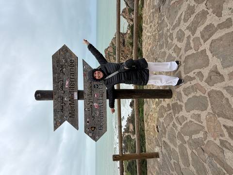 Traveller points happily at a bilingual sign marking the Atlantic and Mediterranean seas with cliff and ocean beyond.