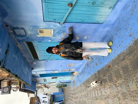 Smiling tourist in bright shoes stands in a blue alley while a curious cat walks by.