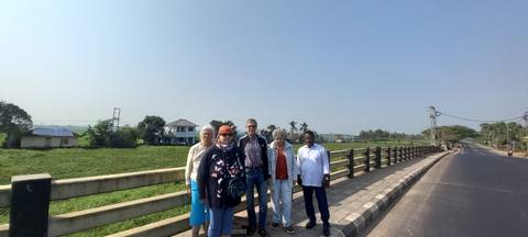 Five travelers pose on a rural bridge with open fields and village houses behind them under a bright sky.
