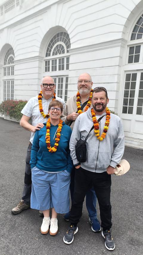 Four travellers wearing marigold garlands posing outside a white colonial building.