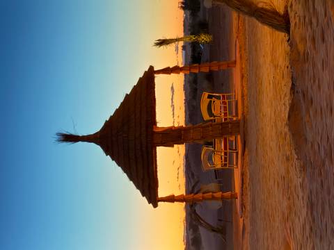 Straw pergola with chairs set on golden desert sand at colorful twilight.