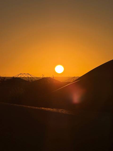 Brilliant orange sun setting behind low desert dunes and silhouetted tent frames, with lens flare in the foreground.