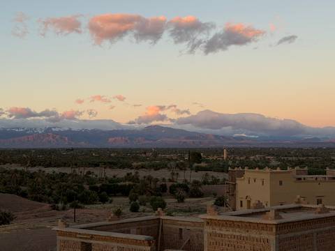 Wide view over a palm-filled valley toward snow-capped mountains glowing pink in the evening light, with an adobe tower in the foreground.