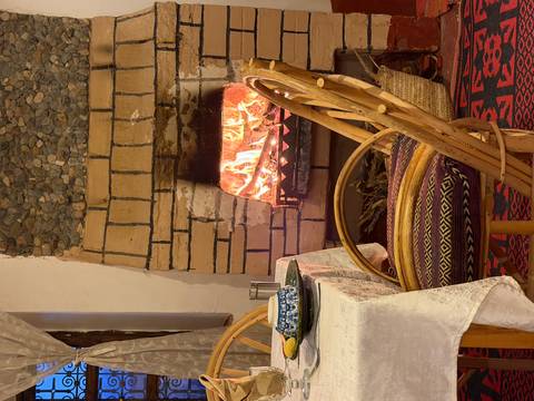 Cozy dining corner with a crackling stone fireplace, set table and woven chairs inside a rustic room.