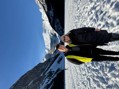 Two smiling tourists pose on the snowy surface of a frozen alpine lake with towering peaks behind.