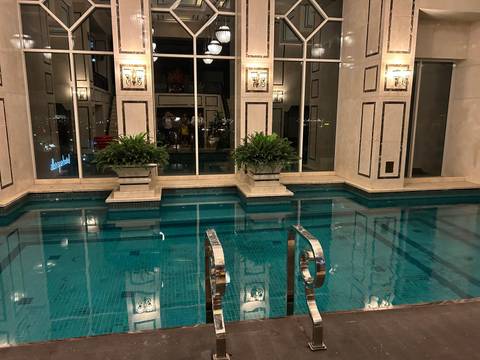 Luxurious indoor swimming pool with turquoise tiles, stainless-steel handrails and potted plants, surrounded by marble walls and tall windows.