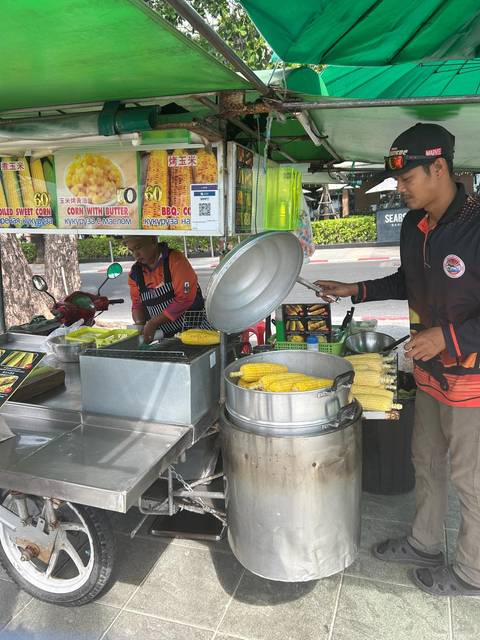 Street food stall where two vendors prepare and sell boiled and grilled corn cobs beside a busy road.