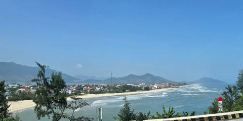 Panoramic coastal view of a long sandy beach, waves, and a small seaside town backed by hills under a clear blue sky.