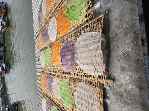 Colorful rice paper sheets drying on woven bamboo racks beside a river dock.