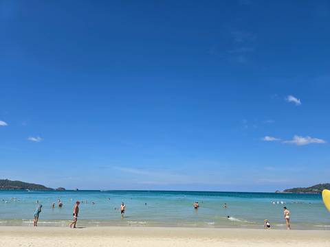 Wide sunny beach with turquoise water and scattered swimmers beneath a vast clear blue sky.