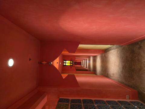 Long narrow corridor painted in warm coral tones with arched doorways and a hanging lantern leading to a distant red door.