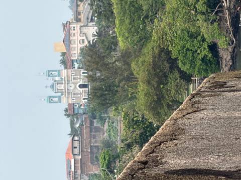 Distant view of a pastel-green church tower rising above lush tropical vegetation and colonial rooftops, seen from atop an old stone fort wall.