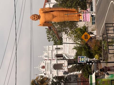 Large roadside orange Buddha statue with a white temple building behind and visitors milling around.