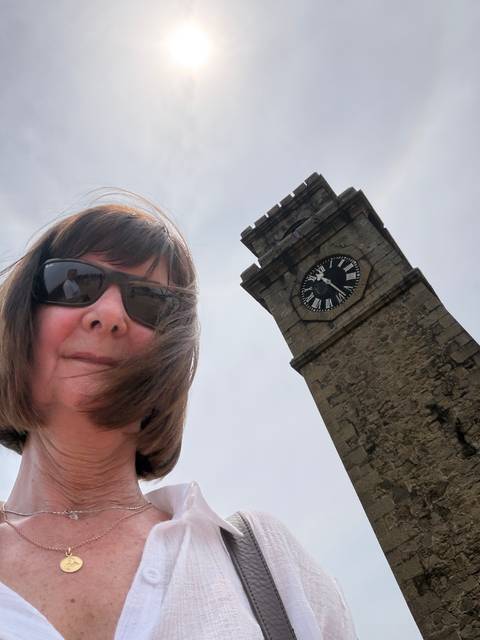 Selfie of a woman wearing sunglasses with the historic Galle Fort clock tower rising behind her against the sky.