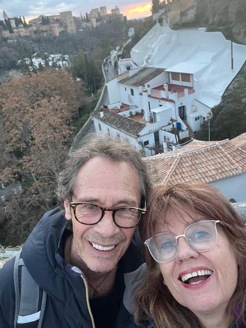 Close selfie of a smiling couple with tiled rooftops and a winding hill road in the background.