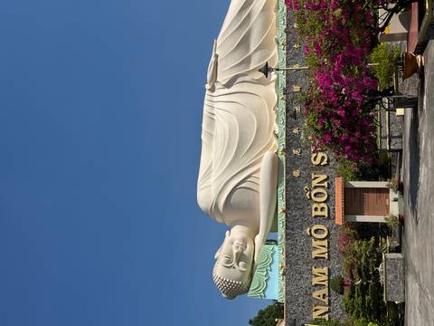 Large reclining Buddha statue on temple rooftop against a clear cobalt sky with blooming bougainvillea.