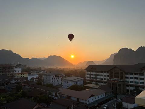 Silhouetted hot-air balloon drifts above misty karst mountains at golden sunrise over a small town.