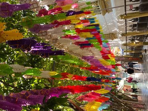 Temple walkway festooned with multicoloured paper lanterns and flags hanging beneath leafy canopy.