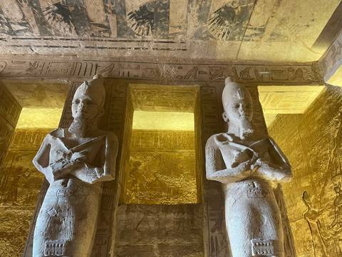 Two towering pharaonic statues stand guard inside a hieroglyph-covered chamber illuminated by warm light.