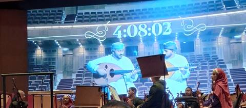 Indoor concert hall scene showing large screen countdown and musicians playing traditional Omani instruments.